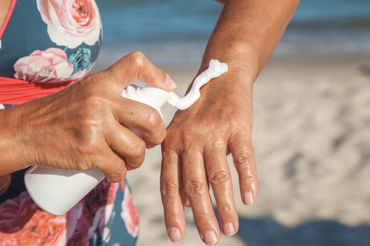 Une femme âgée en maillot de bain frotte ses mains avec un écran solaire dans un conteneur sur la plage de la mer en été