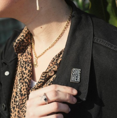 un pin's or et noir sur la veste en jean noire du mannequin, qui a la peau claire et porte une chemise à imprimé léopard.  la police de la broche est en blocs et en majuscules
