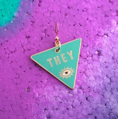 une boucle d'oreille sur un triangle inversé.  le fond est turquoise, et en haut, un oeil et le mot "elles ou ils" en or.